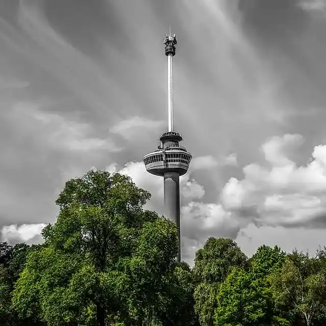 De Euromast in Rotterdam steekt boven de wolken uit, met groene bomen aan de basis en een dramatische mix van sluier- en gewone bewolking.